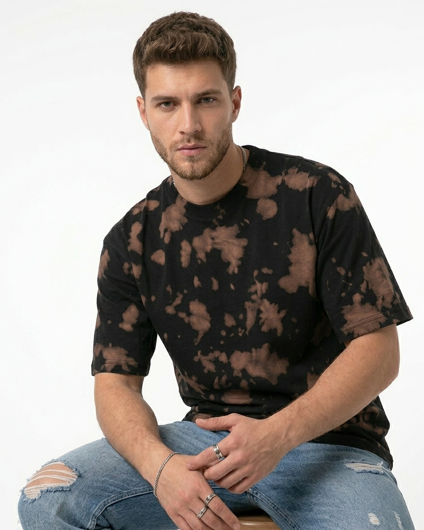 Man wearing a black tie-dye t-shirt and ripped jeans sitting on a stool against a white background